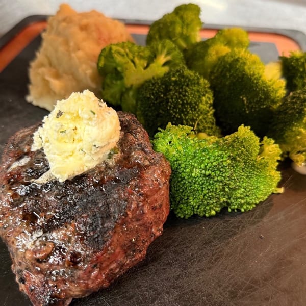 Grilled steak with melting herb butter, mashed potatoes, and steamed broccoli on a dark plate.