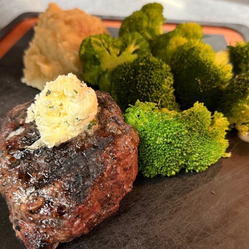Grilled steak with melting herb butter, mashed potatoes, and steamed broccoli on a dark plate.