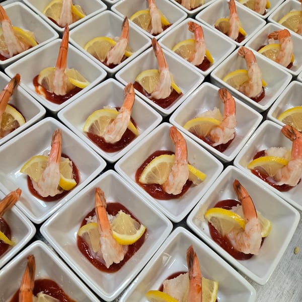 Numerous individual shrimp cocktails in white square dishes with cocktail sauce and lemon wedges.