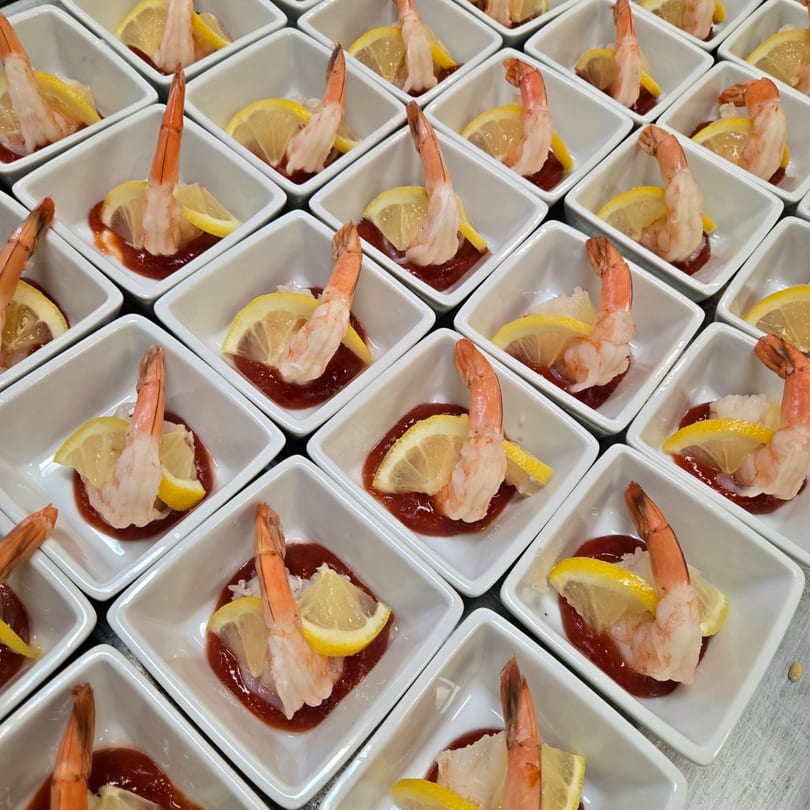 Numerous individual shrimp cocktails in white square dishes with cocktail sauce and lemon wedges.