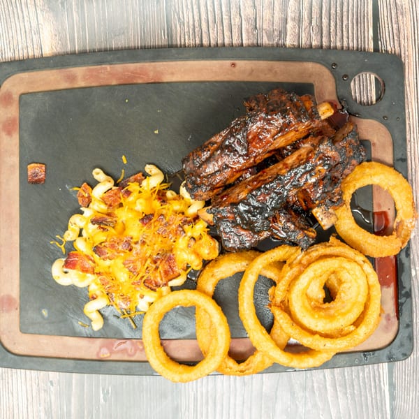 Glazed BBQ ribs, cheesy mac and cheese with bacon, and crispy onion rings on a dark platter.