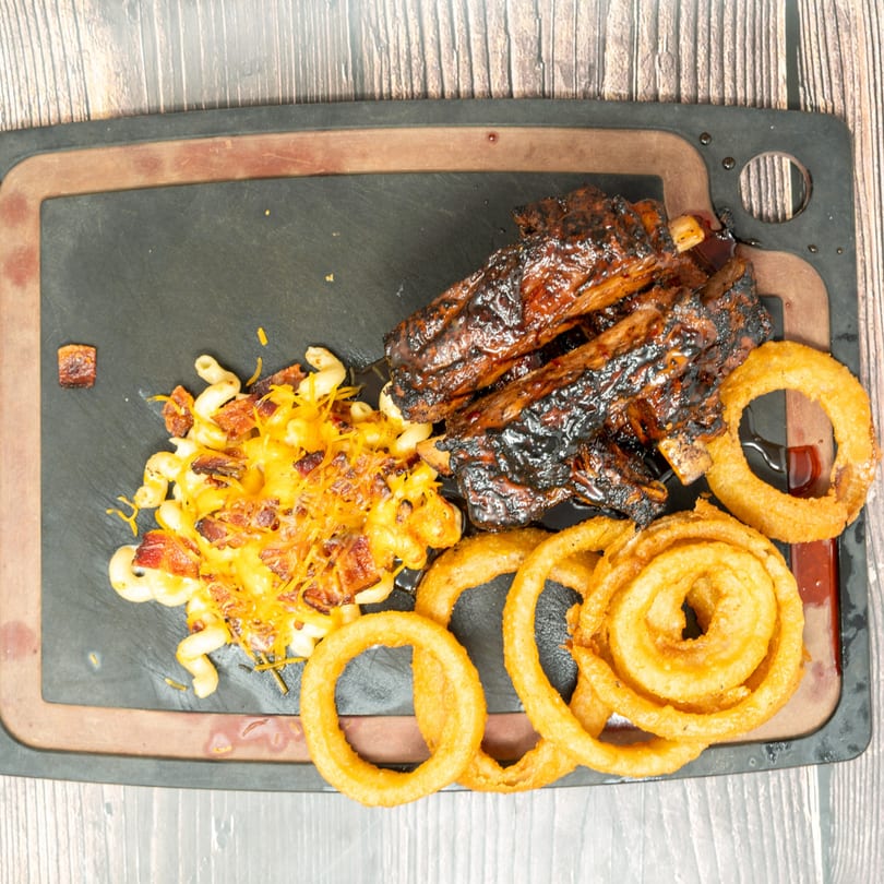 Glazed BBQ ribs, cheesy mac and cheese with bacon, and crispy onion rings on a dark platter.