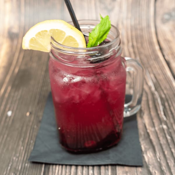 A vibrant red drink in a mason jar with ice, a lemon slice, and mint on a wooden table.