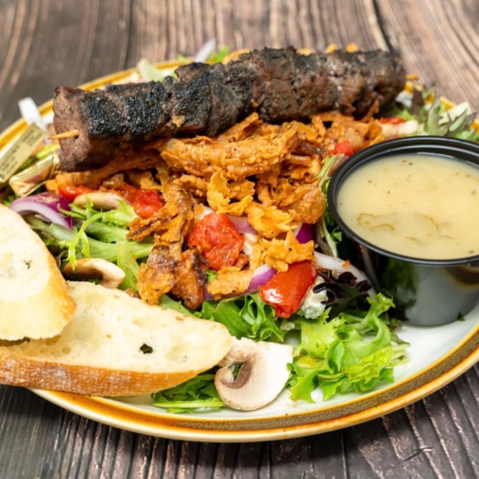Grilled steak skewer on a fresh salad with crispy onions, bread, and a side of dressing.