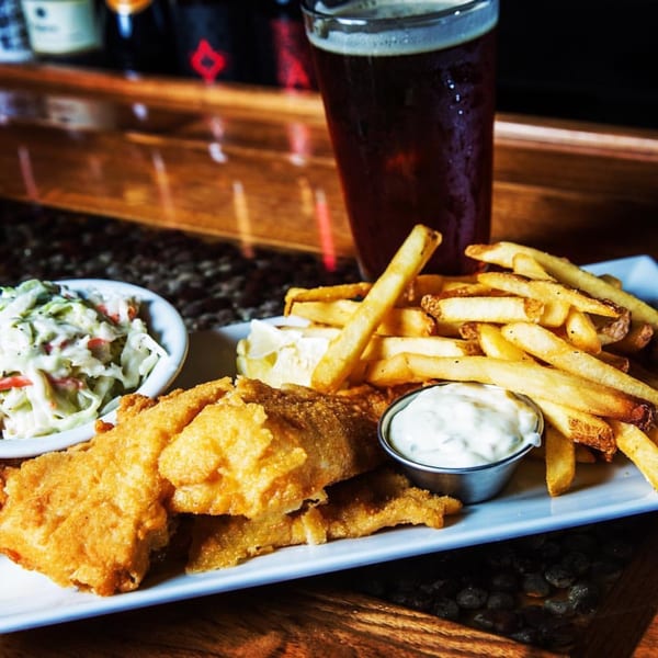 Golden fried fish and chips with coleslaw and tartar sauce, next to a dark beer on a wooden bar.