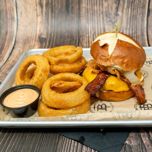 A pretzel bun burger with bacon, cheese, and onions, served with onion rings and dipping sauce.