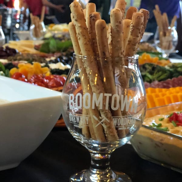 Boomtown Brewery glass filled with breadsticks, surrounded by a buffet of appetizers and dips.