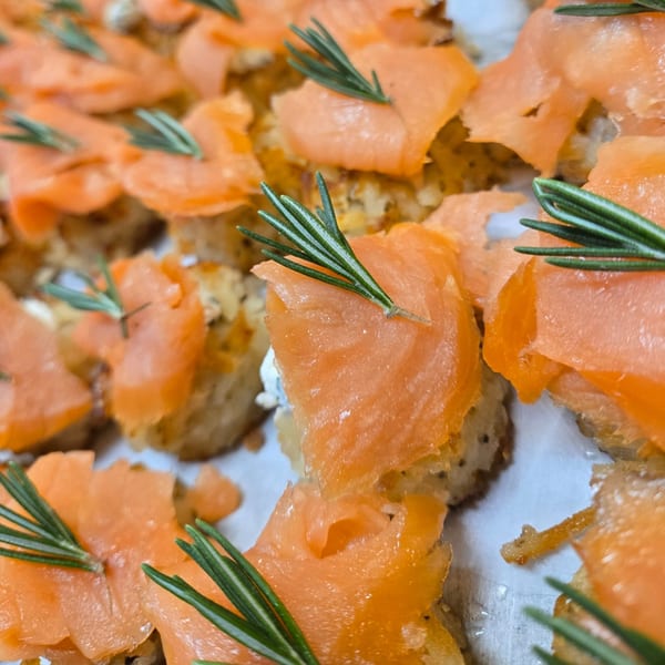 Close-up of numerous smoked salmon canapés with cream cheese and rosemary sprigs on a white platter.