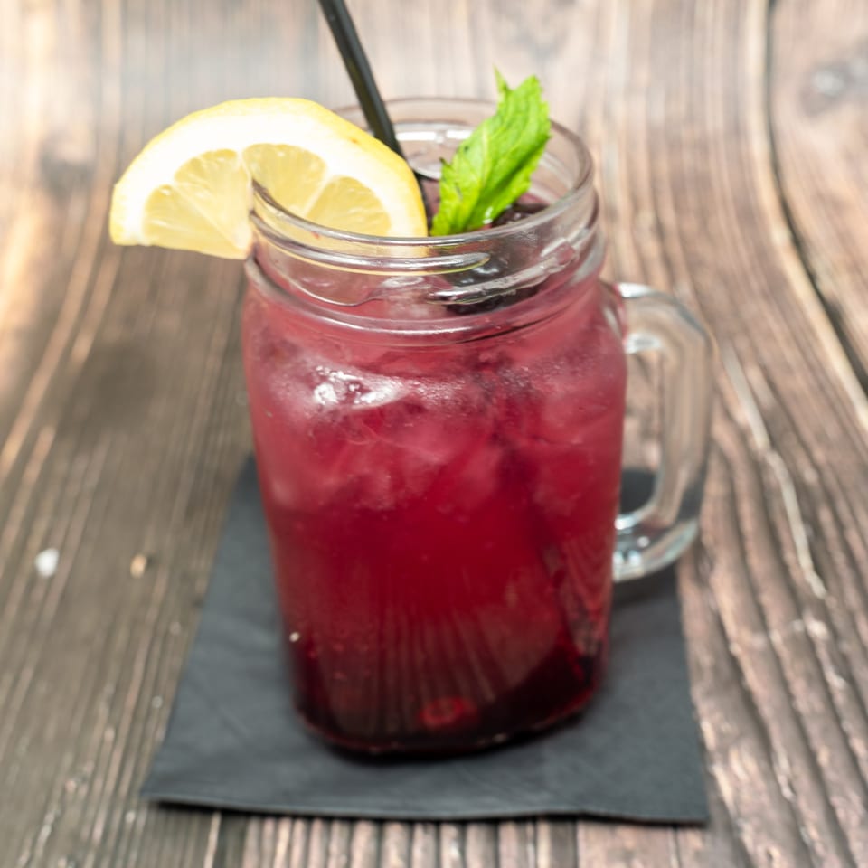 A vibrant red berry cocktail in a mason jar, garnished with a lemon slice and mint.