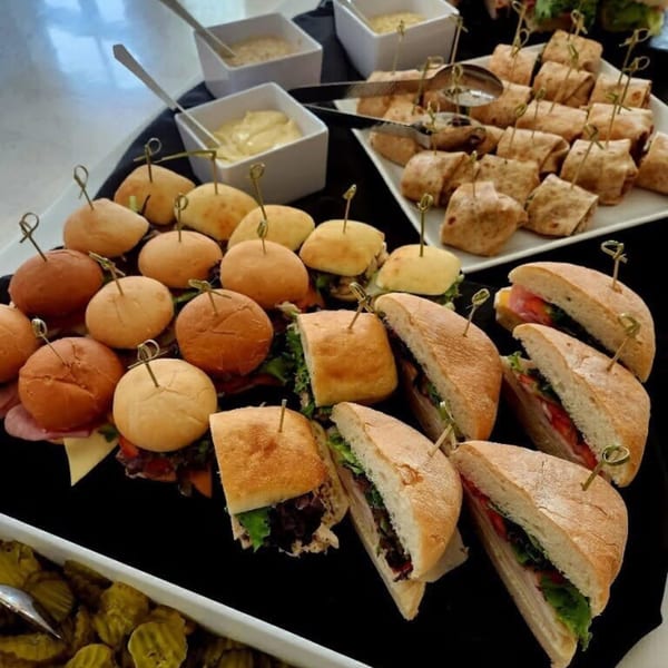 A catering spread with mini sandwiches, sliders, wraps, and bowls of sauces and pickles.