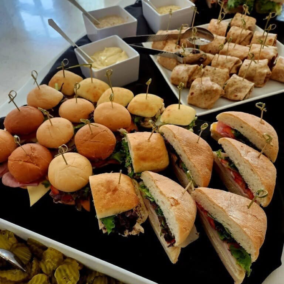 A catering spread with mini sandwiches, sliders, wraps, and bowls of sauces and pickles.