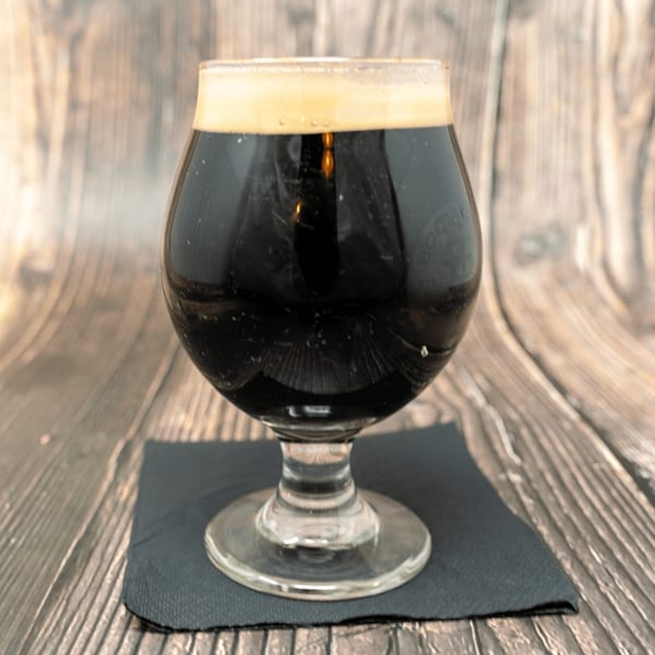 Dark stout beer with a frothy head in a snifter glass on a black napkin.