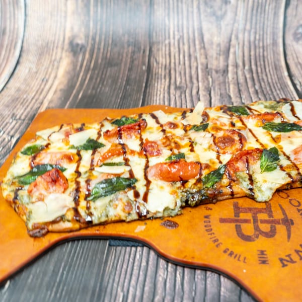A rectangular flatbread pizza with melted cheese, tomatoes, basil, and a balsamic glaze drizzle.