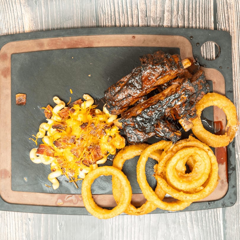 BBQ ribs, cheesy macaroni, and crispy onion rings on a dark serving board.