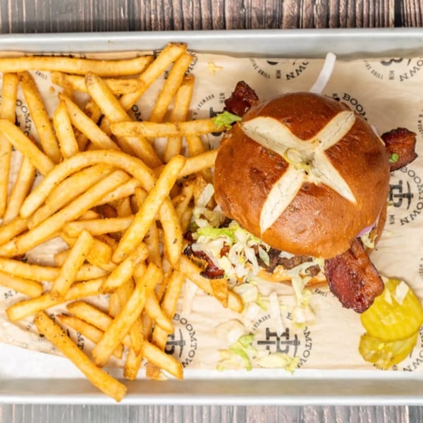 A pretzel bun burger with bacon, lettuce, and pickles, served with a side of fries.