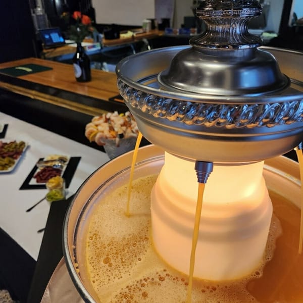 A beverage fountain dispenses orange liquid into a bowl, with appetizers and a bar in the background.